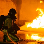 links kniet ein Feuerwehrmann in Schutzkleidung und l&ouml;scht einen Brandherd am Boden