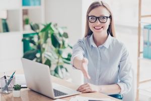 Junge Frau am Schreibtisch streckt dem Betrachter zur Begr&uuml;&szlig;ung die Hand hin