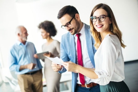 Mann und Frau in Businesskleidung im Gespr&auml;ch, Frau wendet sich zum Betrachter, im Hintergrund zwei weitere Personen